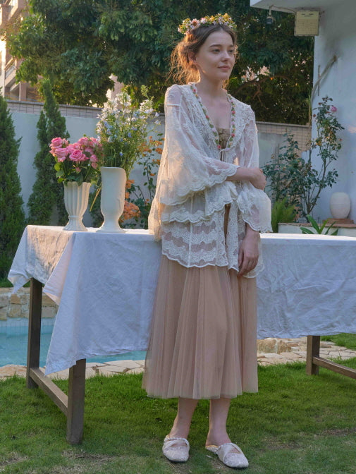 A woman wearing a layered lace cape and nude tulle skirt stands beside a floral garden table, styled in an elegant cottagecore outfit.