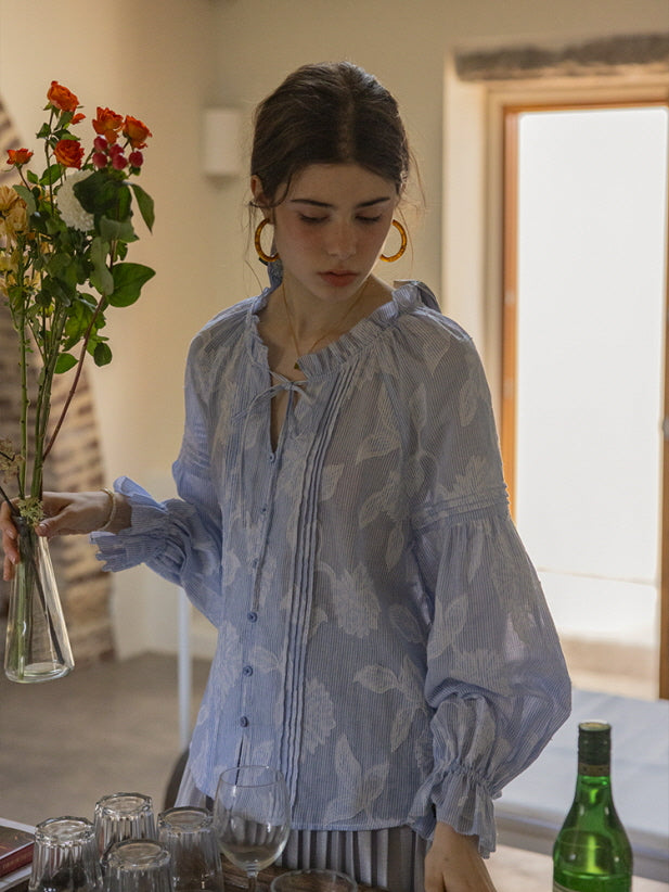 A young woman holding a vase of roses while wearing a blue floral blouse with puffed sleeves and delicate ruffle details. The warm, airy setting complements the dreamy, fairytale-inspired look.