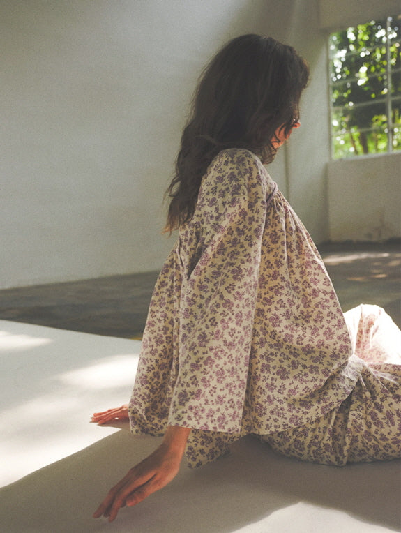 The model relaxes on the floor in soft light, wearing a vintage-style lavender floral loungewear set that flows gently with a dreamy silhouette.