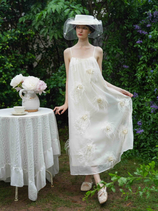 Standing beside a tea table, the model wears a cream dress with feather flower accents, styled for a romantic fairycore-inspired outdoor scene.