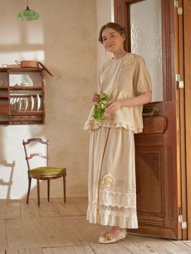 A woman in a soft beige blouse and embroidered skirt holds a floral wreath in a rustic room, evoking a serene cottagecore afternoon.
