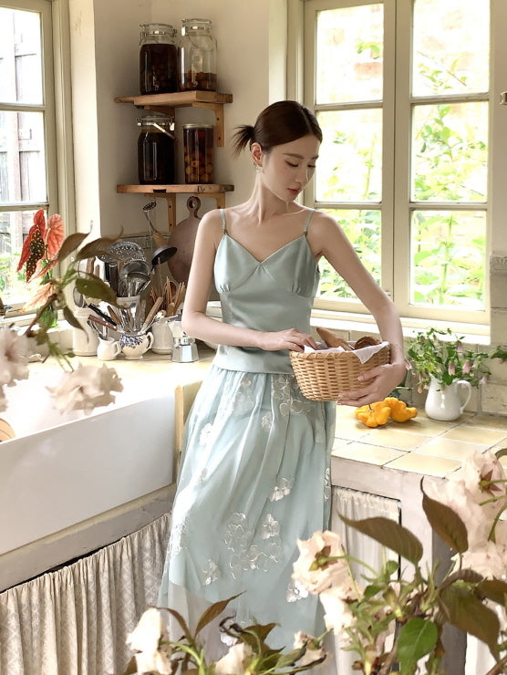 Wearing a light green satin and tulle fairy skirt and camisole, a woman gently holds a basket of bread by the window, surrounded by flowers and vintage kitchen decor.