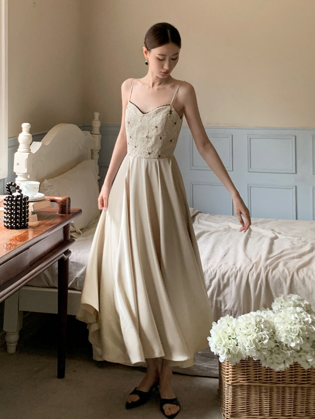 A young woman stands gracefully by a vintage bed in a soft beige fairycore dress with lace bodice and flowy skirt, evoking a romantic mood.