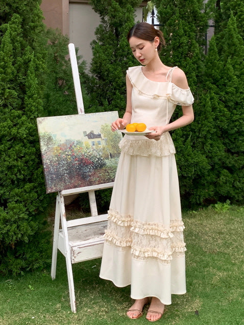 Wearing an off-shoulder cream top with ruffles and a tiered lace-trim skirt, the model stands in a garden with a painting and plate of lemons.