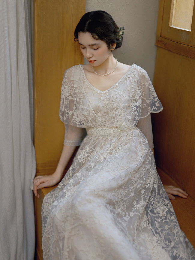 Seated in a quiet corner, a woman wears a romantic ivory gown with floral lace and pearl details, evoking a soft cottagecore aesthetic.