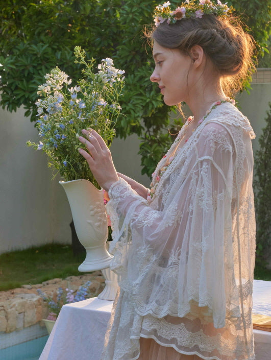 A woman in a layered lace robe and floral crown gently touches a vase of wildflowers, styled in a soft fairycore garden atmosphere.