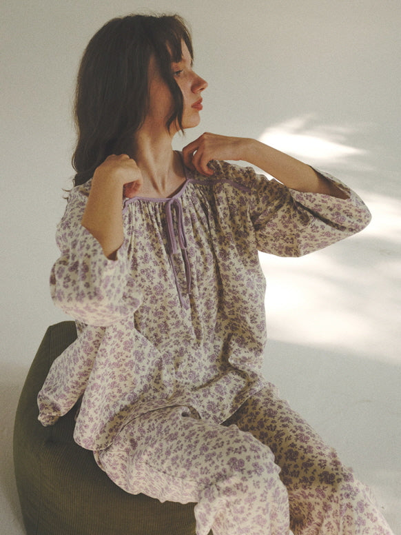 Sitting calmly in morning light, the model adjusts the ribbon neckline of a purple floral pajama set, capturing romantic vintage loungewear charm.