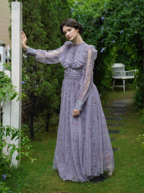 Wearing a sheer lavender fairycore dress, the model poses thoughtfully in a garden path, evoking vintage elegance and soft charm.