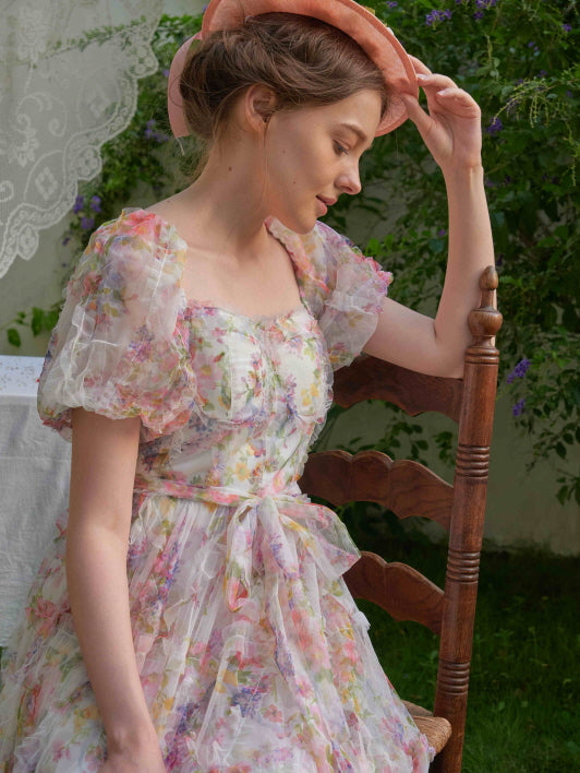 The woman gently adjusts her bonnet while sitting in a garden, wearing a pastel floral dress with puff sleeves and layered sheer fabric.