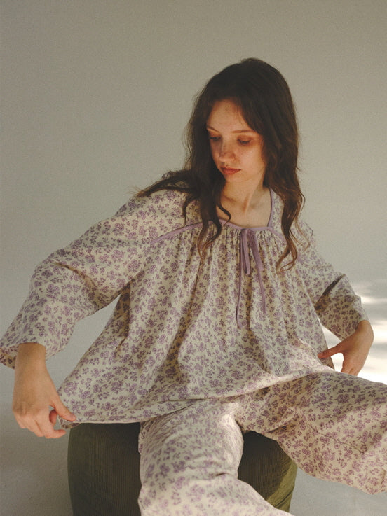 The model gently pulls at the hem of a loose floral loungewear set, wearing relaxed cottagecore pajamas with a calm, dreamy expression.