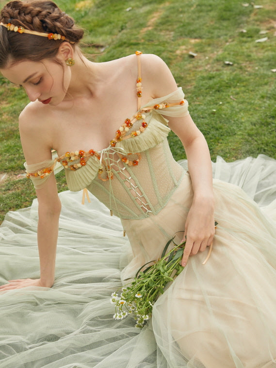 A woman sits gracefully on grass in a fairytale-inspired green corset dress adorned with orange floral accents and soft tulle layers.