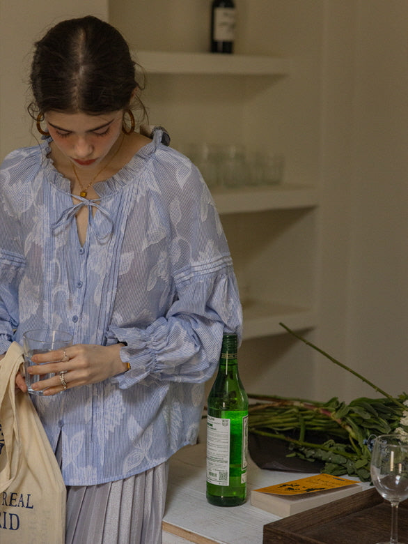 A young woman in a blue and white floral blouse with ruffled cuffs and a tie neckline, standing by a kitchen counter while holding a glass and a canvas tote. The soft, cottagecore style exudes a relaxed, vintage-inspired aesthetic.