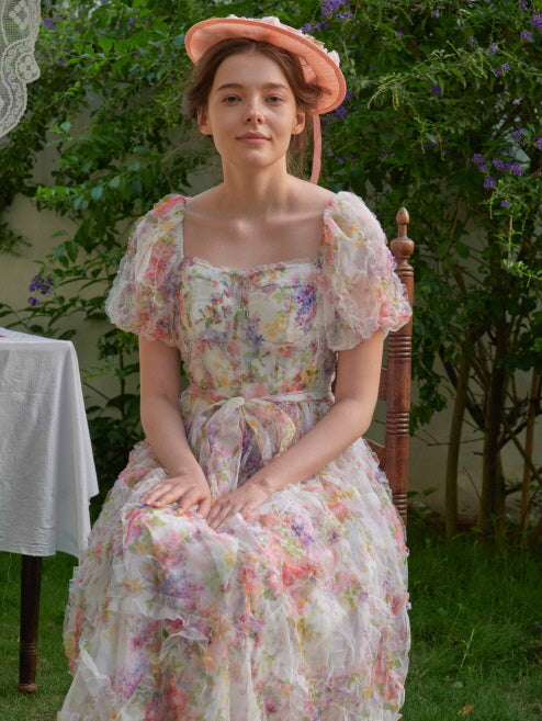 A woman in a pastel floral fairycore dress sits gracefully on a vintage chair, surrounded by romantic greenery and soft lace décor.