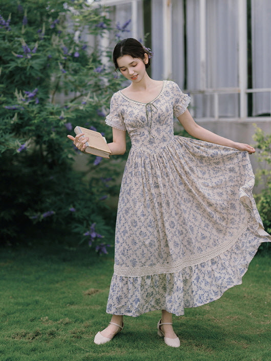 A graceful, vintage-style dress with a blue floral pattern and lace trim, as the model gently lifts the hem while holding a book, evoking a fairytale-inspired moment.