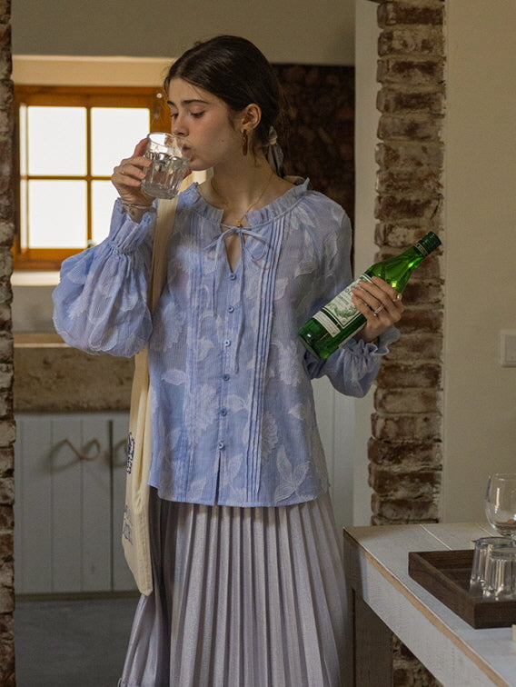A young woman sipping water while holding a green glass bottle, wearing a blue floral blouse with puffed sleeves and a pleated skirt. The relaxed, fairytale-inspired aesthetic creates a cozy, vintage feel.