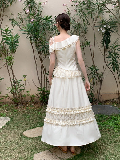 The model showcases the back view of a cream one-shoulder top and tiered lace skirt while walking through a garden in a fairytale-inspired look.