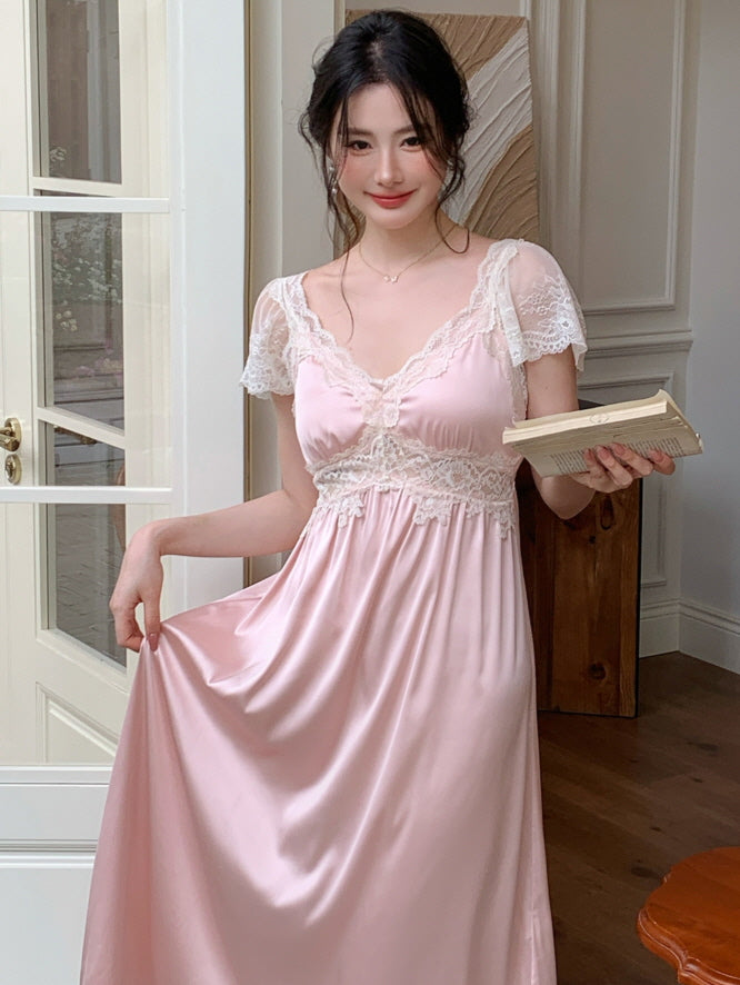 A young woman stands by a glass door in a pastel pink lace princess nightgown, smiling softly while holding an open book. Her look evokes a soft, vintage cottagecore aesthetic.