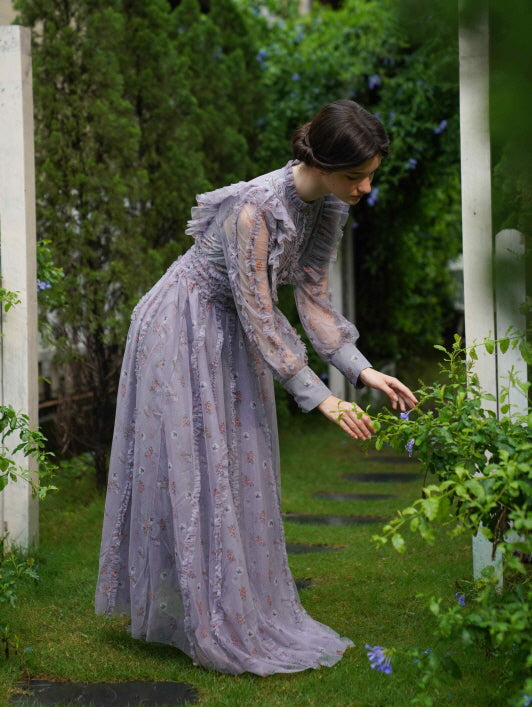 In a romantic lavender dress, the model leans toward blooming flowers along a garden path, expressing a serene and dreamy fairycore mood.