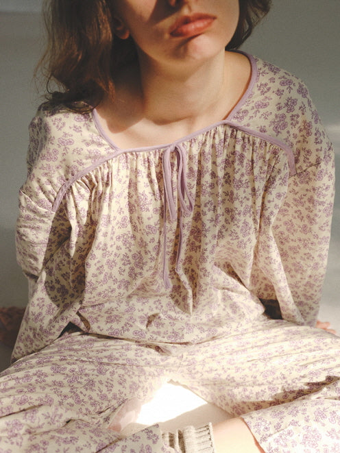 The model sits quietly in soft lavender floral loungewear, sunlight highlighting the gathered neckline and evoking gentle cottagecore comfort.