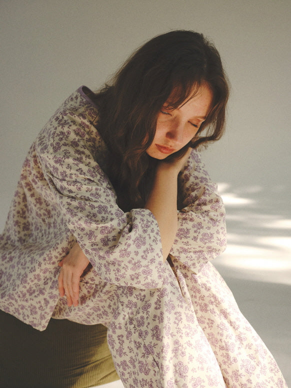 Resting her head on her knees, the model wears a relaxed purple floral pajama set, radiating a calm, introspective cottagecore atmosphere.
