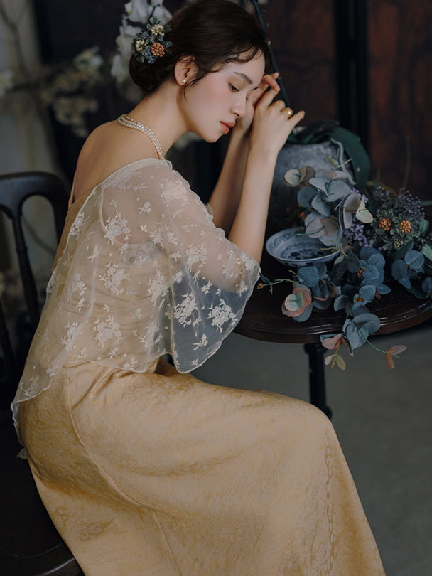 Dressed in an elegant gold gown with sheer embroidered overlay, a woman stands near flowing curtains, evoking a refined princesscore mood.