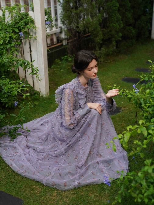 The model sits on the grass in a flowing lavender floral dress, gently reaching toward a bloom in a dreamy fairycore garden setting.