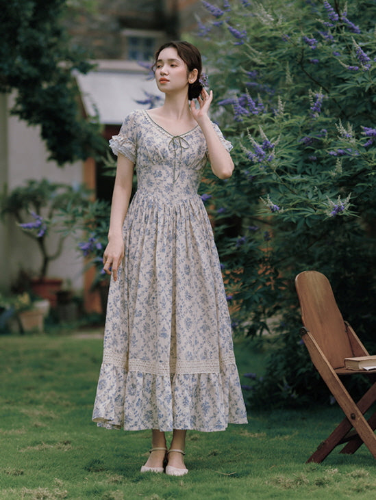 The model stands serenely in a garden setting, wearing a romantic, cottagecore floral dress with puff sleeves and a lace-up neckline, exuding timeless elegance.