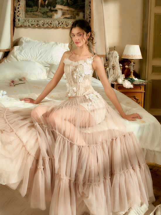 The model sits elegantly on a romantic bed, wearing a blush floral corset and sheer ruffled tulle skirt, evoking a soft vintage fantasy mood.