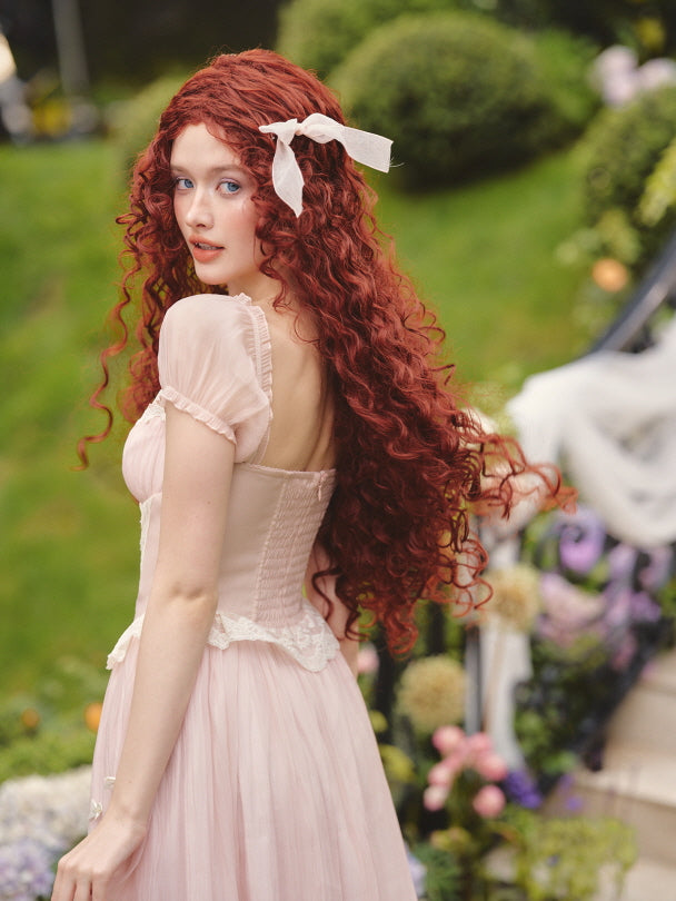 The model turns back with flowing red curls while wearing a pink lace corset dress, evoking a dreamy fairytale aesthetic in a garden scene.