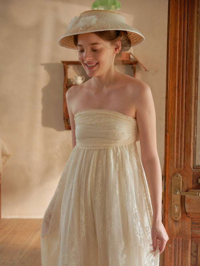 A young woman smiles softly in a strapless lace ivory dress, evoking a dreamy vintage mood in a cozy cottagecore setting.