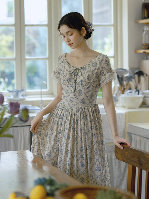 A soft, vintage-inspired floral dress with lace accents and a delicate bow-tie neckline, as the model stands in a sunlit kitchen, creating a dreamy cottagecore ambiance.