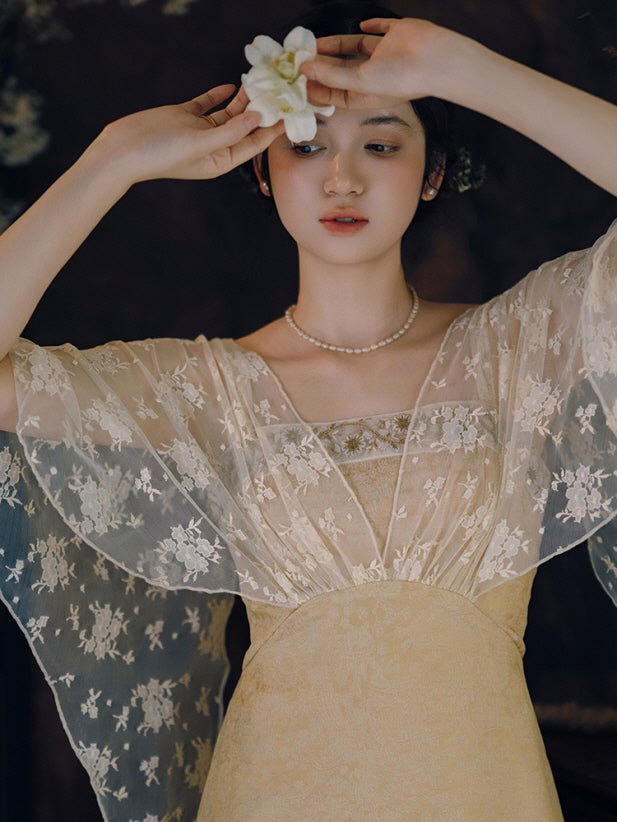 A woman gently raises her arms while holding a white blossom, wearing a soft gold dress with sheer floral sleeves in a fairycore mood.