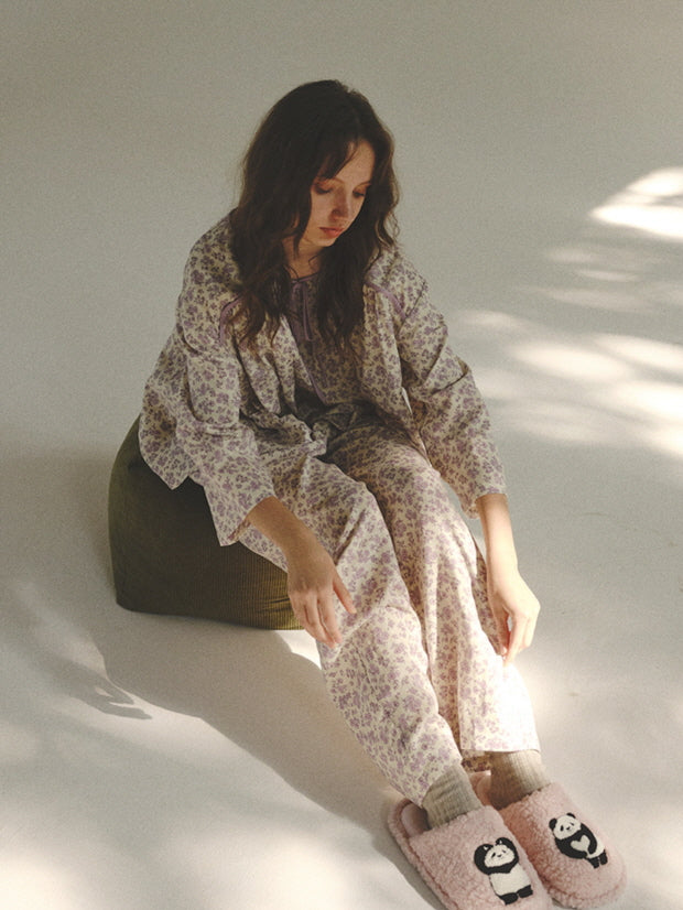 Sitting on a green pouf in cozy pink slippers, the model wears a romantic floral loungewear set, capturing a soft and dreamy cottagecore moment.