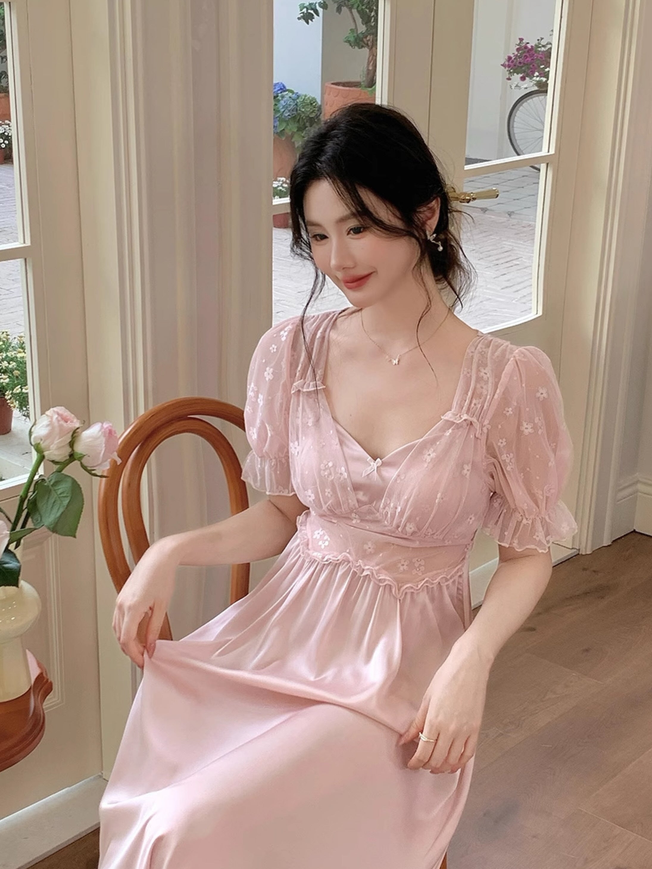 Model sitting beside a window in a blush pink satin nightgown with sheer floral puff sleeves, styled with romantic coquette elegance.