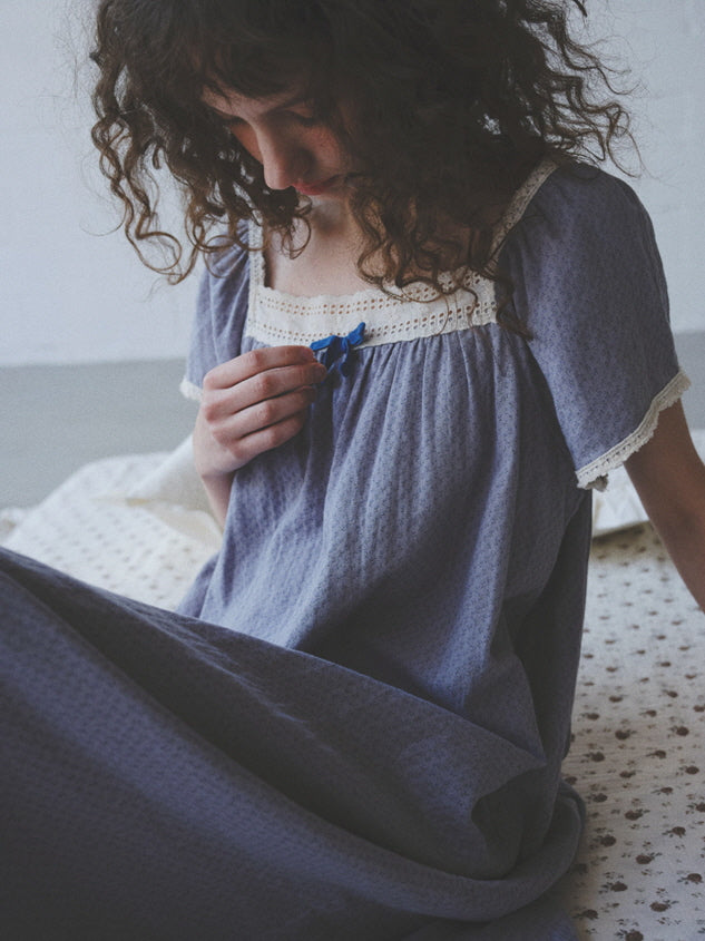Wearing a flowy lavender nightgown with a lace square neckline and blue ribbon, the model stands in gentle light for a dreamy cottagecore look.
