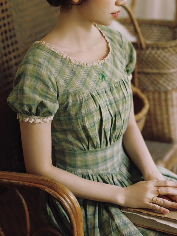 A back view of a woman in a green plaid dress with a waist tie, leaning over a vintage-style kitchen sink adorned with hydrangeas and classic ceramic decor. The scene captures a nostalgic, fairytale-inspired cottagecore moment.
