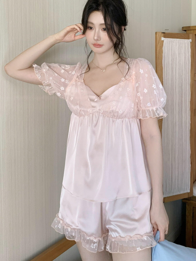 Model wearing a silky pink fairycore pajama set with floral mesh sleeves and ruffled hem, standing calmly in a softly lit room with wooden decor.