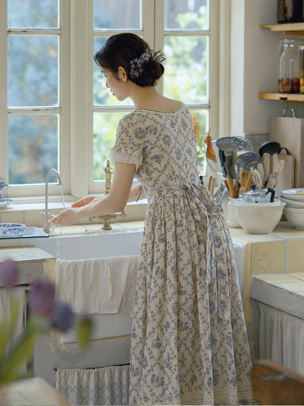 Back view of a romantic, cottagecore dress adorned with blue floral prints and a ribbon-tied waist, as the model gently washes dishes in a quaint, sunlit kitchen.