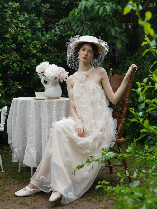 Model in a flowing beige tulle dress with floral details and ribbon neckline sits gracefully in a garden, capturing a vintage-inspired fairytale mood.