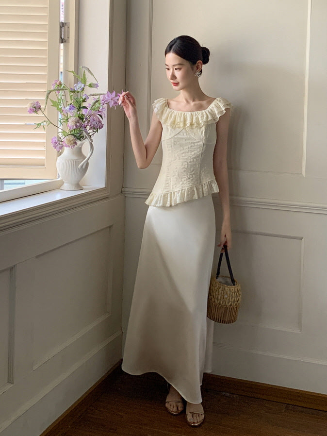 A woman stands by a vintage window in a cream ruffled top and satin skirt, holding a basket bag in a dreamy fairycore setting.