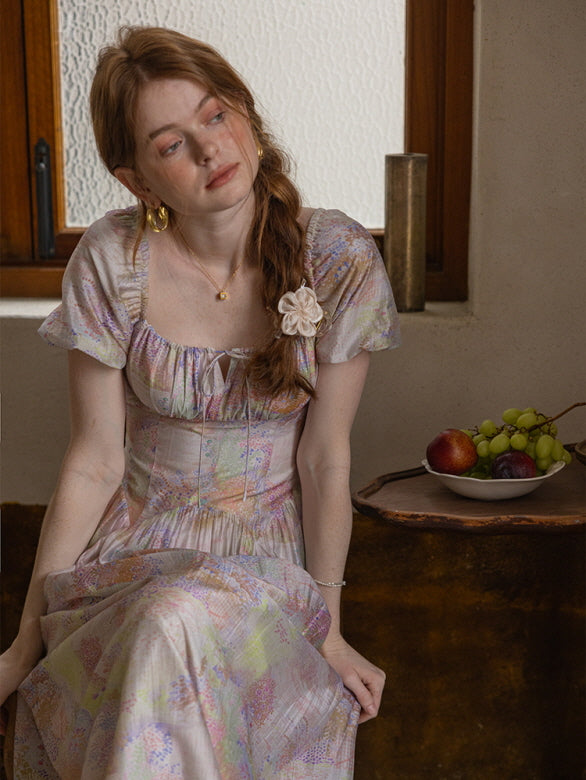 A woman sits by a rustic window, wearing a pastel floral dress with puff sleeves and a lace-up neckline, exuding a serene, vintage-inspired look.