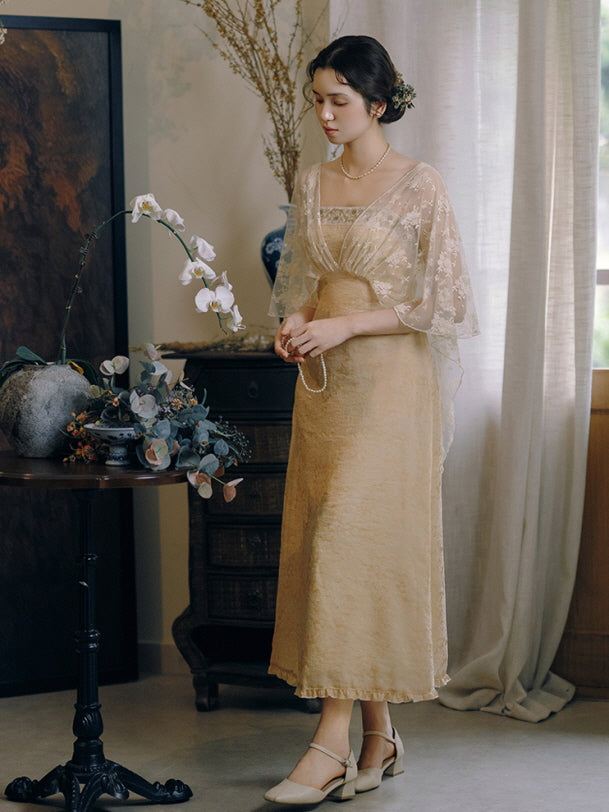In a serene vintage room, a woman in a golden jacquard gown with lace overlay stands beside a table of orchids, evoking a romantic aesthetic.