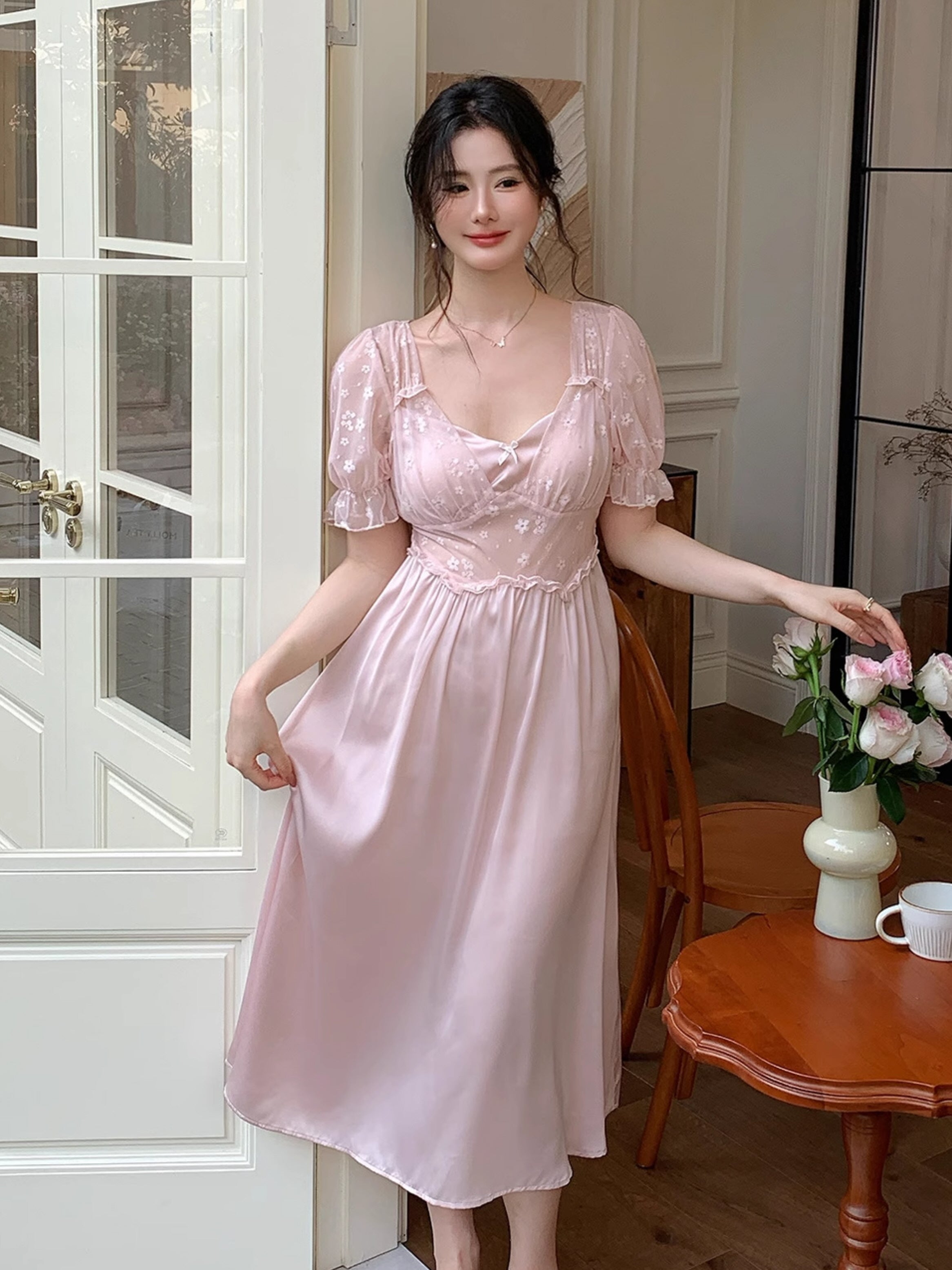 Woman standing by a café table in a princesscore loungewear dress with a fitted mesh bodice and soft satin skirt in pastel pink.