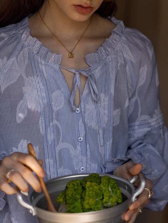 Close-up of a young woman wearing a blue floral blouse with a ruffled neckline and tie detail, stirring fresh greens in a pot. The vintage-inspired look adds a soft, cottagecore charm.