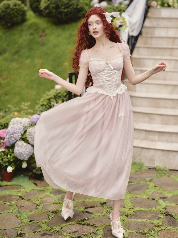 Wearing a princesscore-inspired pink dress with puff sleeves and corset lace-up detail, the model poses gracefully on a garden path.