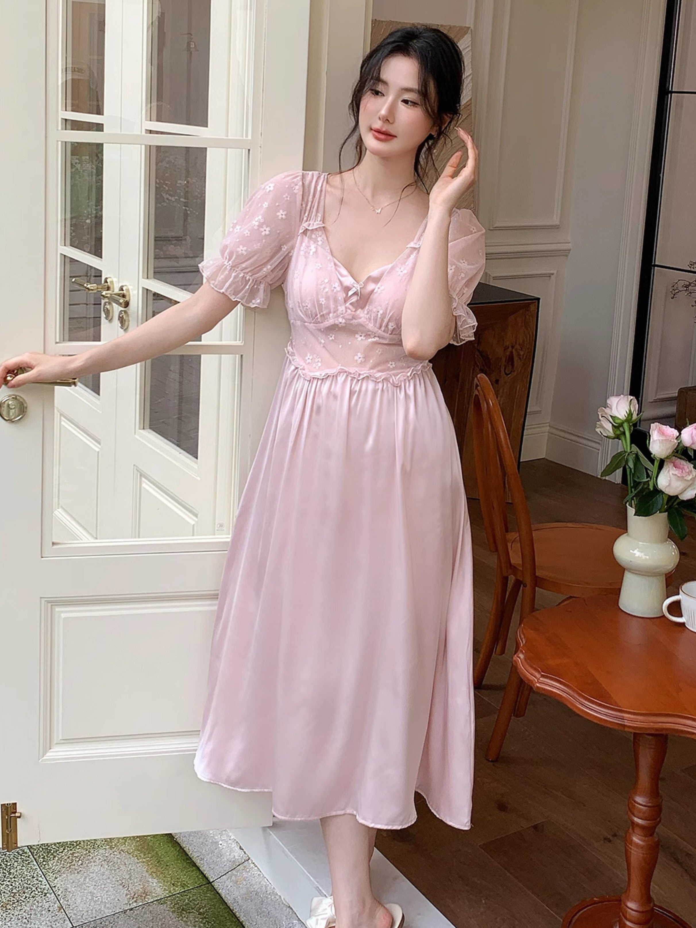 Model gently touching her hair in a soft pink fairytale nightdress with embroidered mesh sleeves, standing by a sunlit garden door.
