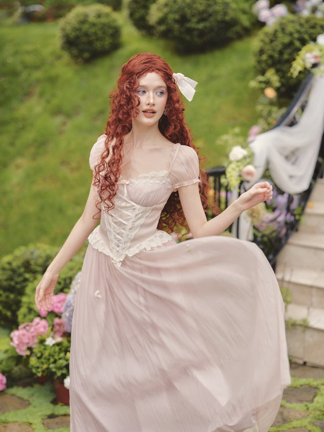 The model twirls in a soft pink fairytale dress with puff sleeves and a lace-up corset bodice, surrounded by blooming flowers in a garden setting.