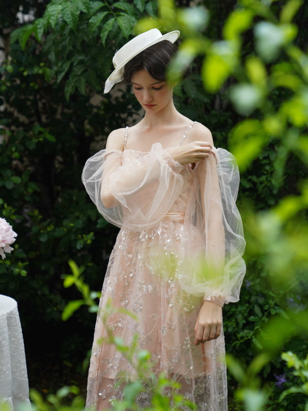 Model adjusts her sheer puff sleeves in a sparkling blush dress, surrounded by greenery for an ethereal fairycore and princesscore moment.