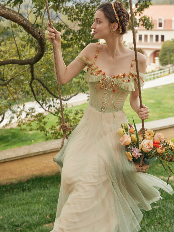 Model sitting on a rustic swing holding a flower basket, wearing a vintage-inspired green corset fairy dress with floral accents and a dreamy silhouette.
