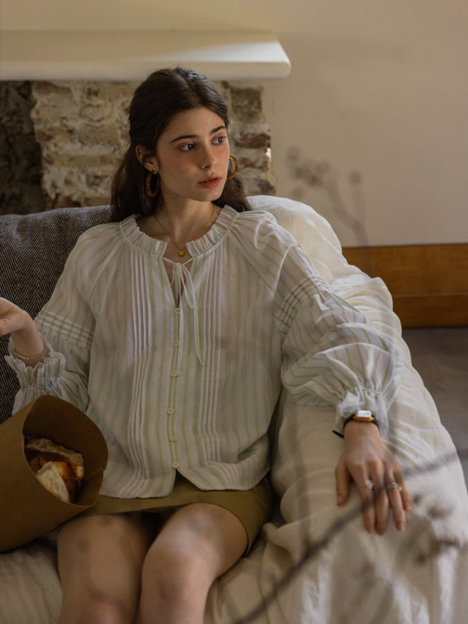 A young woman seated on a cozy sofa, wearing a light green and white striped blouse with ruffled cuffs and a tie neckline, holding a paper bag of pastries. The soft, cottagecore aesthetic creates a warm, vintage-inspired atmosphere.